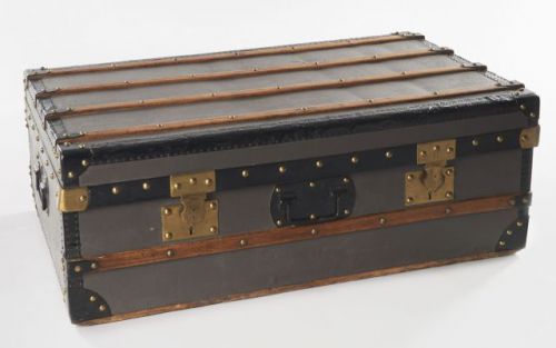 ANONYME, Malle de voyage en toile grise sur bois, bordures en métal noir clouté et laiton aux angles. Lattes de renfort en bois clouté. Intérieur en tissu rouge à carreaux. Deux fermoirs en laiton. Deux poignées de portage en métal sur les côtés. Restaura